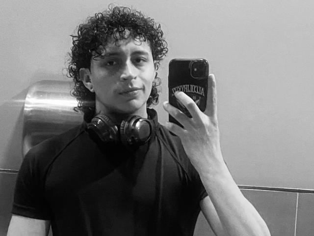 JayTaylor, avec ses cheveux bouclés, portant une chemise noire, tenant un téléphone dans un autoportrait pris dans un miroir, a des écouteurs autour du cou, et regarde légèrement sur le côté avec un sourire discret..
