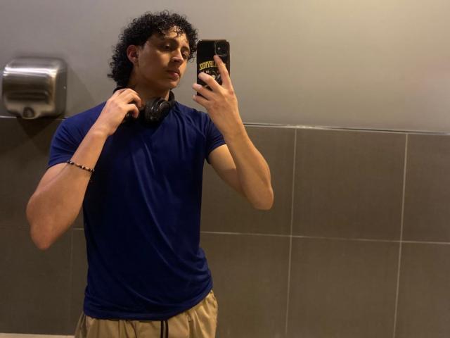 JayTaylor, avec ses cheveux noirs bouclés, un t-shirt bleu, un pantalon beige, tenant des écouteurs noirs autour du cou, prenant un selfie devant un miroir avec son téléphone, portant un bracelet noir, dans une salle de bain avec un mur en carreaux gris..