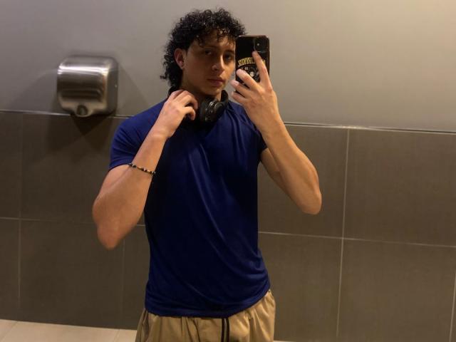JayTaylor, avec ses cheveux noirs bouclés, portant un t-shirt bleu et un pantalon beige, tenant des écouteurs près de son cou, prenant un selfie devant un miroir dans une salle de bain..