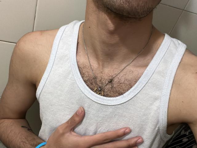 MiloJhones wearing a white tank top, chest covered in dark hair, silver necklace, hand on chest, short beard, light skin, visible tattoo on left arm.