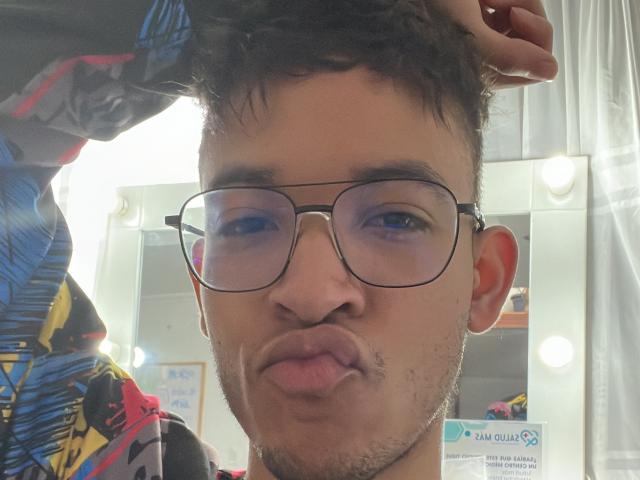 MiloJhones, with glasses, puffy lips, and a slight beard, has his hand in his messy brown hair. His face shows small scratches. He's wearing a colorful, patterned shirt.