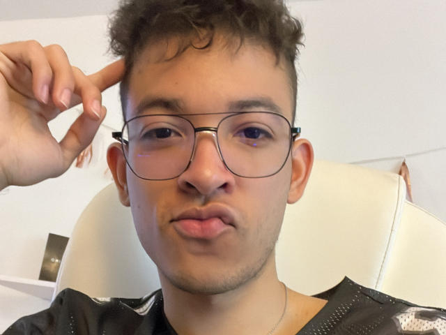 MiloJhones with curly brown hair, glasses, and a slightly pouty face, sticking his tongue out while pointing to his left temple. Wearing a black shirt with white patterns.