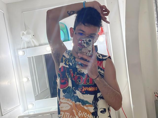 MiloJhones with short black hair, wearing a colorful sleeveless shirt, holding a phone with a sticker-covered case, taking a mirror selfie, arm raised, white bathroom background.