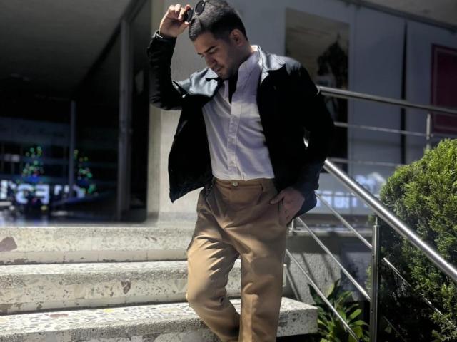 TonyLatino with dark hair, black leather jacket, white shirt, tan pants, standing on concrete stairs, hand in pocket, other hand touching head, metal railing, sunlight, bush in foreground.