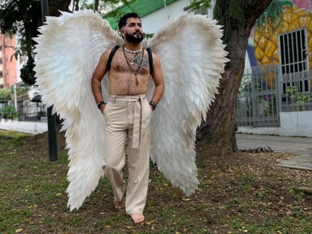 TonyLatino with huge white angel wings, shirtless, hairy chest, beige pants, black backpack, bearded, standing in grass, wearing multiple necklaces, barefoot, brown skin, looking up.
