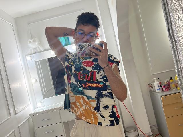 MiloJhones with curly dark hair, blue glasses, and a colorful graphic shirt, taking a mirror selfie. He's holding a phone with a reflective case, white drawers behind.