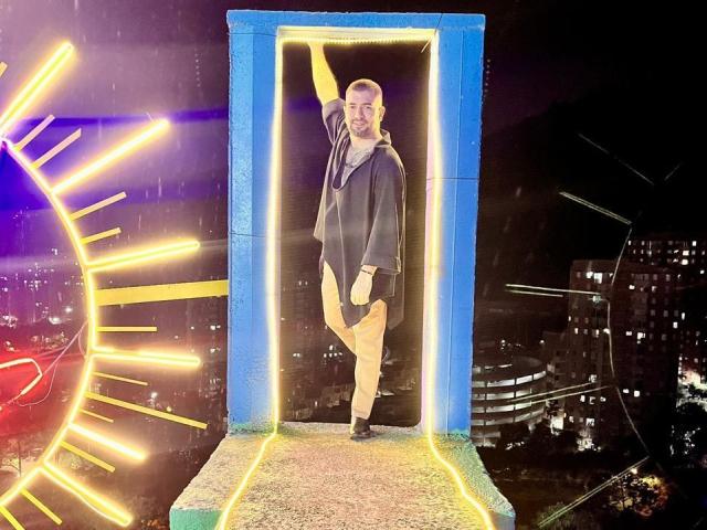 TonyLatino stands in a blue-lit doorway, wearing a black hoodie and tan pants, with neon yellow lights on the left and city lights in the background.