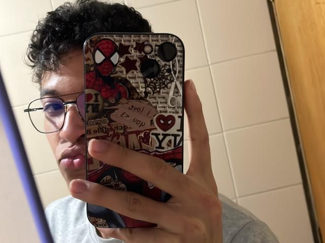 MiloJhones with curly black hair and glasses, holding a phone with a Spider-Man sticker case, taking a mirror selfie in a bathroom. His hand is covering his ass.