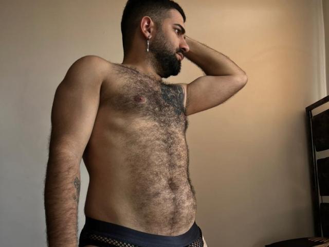 TonyLatino, shirtless, with a hairy chest and tattoos, wearing black mesh underwear, has a beard, ear ring, and is looking to the side with one hand on his head.