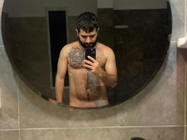 TonyLatino, shirtless with a beard, dark hair, tattoo on right shoulder, holding phone in mirror selfie, chest and belly hair, standing in bathroom, taking mirror photo.