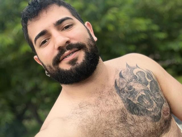 TonyLatino, shirtless with a beard, dark curly hair, and a hairy chest, has a detailed wolf tattoo on his right pectoral. Smiling, he's outdoors with greenery in the background.