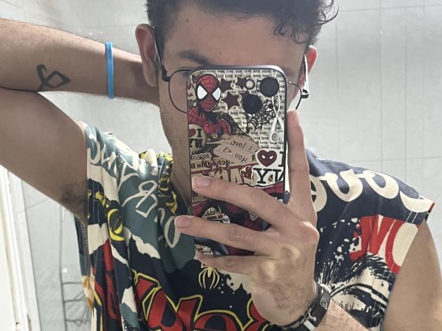 MiloJhones taking a mirror selfie, wearing a colorful graphic tee, glasses, and a blue bracelet, with a phone case featuring Spider-Man and street art. His arm has a heart tattoo.