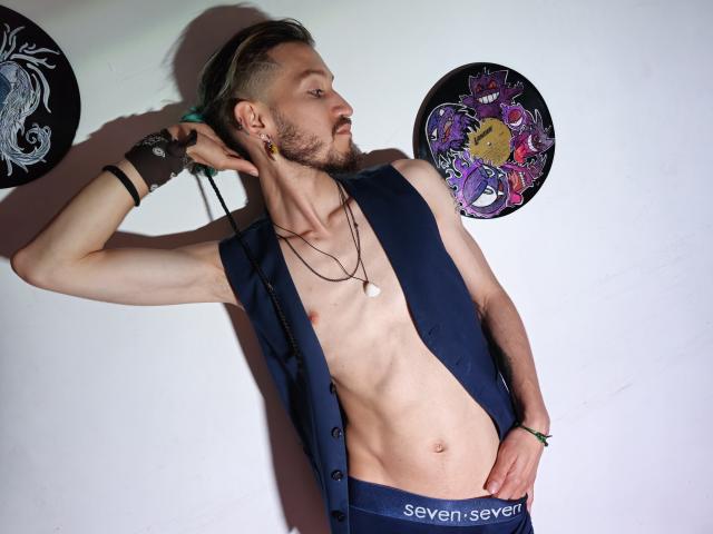 AngelDamianArt, sin camisa con un chaleco azul, abdominales tonificados, mano en el pantalón, tapones para los oídos, pulsera, collar, cabello corto castaño, perfil lateral, mirando a la izquierda, tatuajes en la pared, de pie, con confianza..