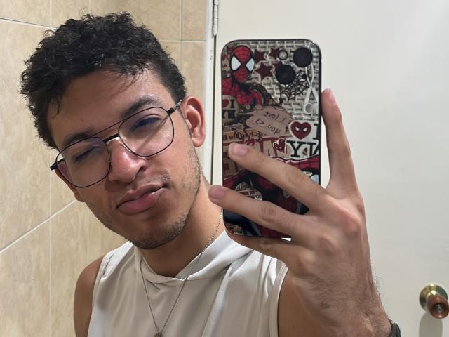 MiloJhones with curly black hair, glasses, and a white sleeveless shirt, pouting lips, holding a phone with a colorful case, in a bathroom. His ass is not visible.