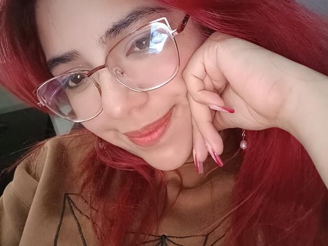 RousseColliinss with red hair, glasses, and pink nails, smiling with hand on cheek, brown top, reflection in glasses, and subtle makeup, showing cute, sexy face.