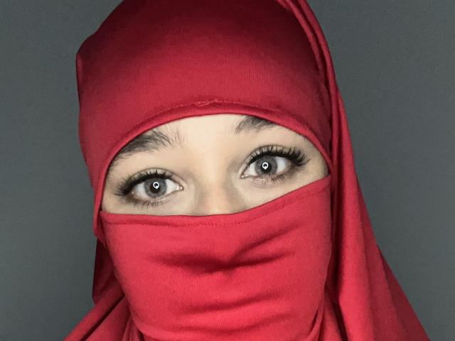 RaishaX has big brown eyes, thick black eyebrows, and is wearing a red headscarf and mask, looking straight into the camera with a serious expression.
