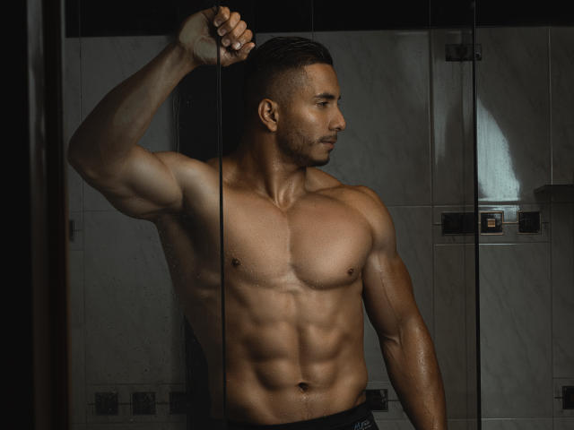 ZaneMaverick, shirtless, muscular, wet, standing in a dark, tiled shower, holding a glass door with a raised arm, showcasing a strong, defined, tan, sexy chest and abs.