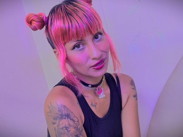 MilaRouge with pink and black hair in double buns, black tank top, pink lipstick, choker with pink gem, tattoos on arm, smiling, purple and pink lighting.