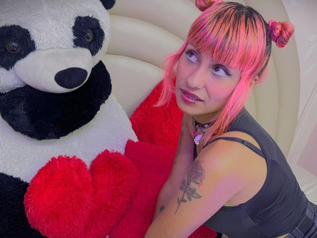 MilaRouge with pink and orange hair in pigtails, wearing a black tank top, sitting next to a giant panda stuffed toy with red heart-shaped bottoms. Tattoo on her arm.