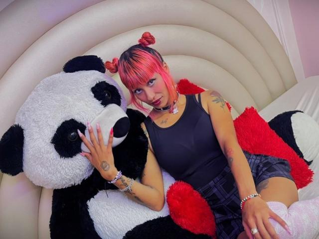 MilaRouge with pink hair in buns, black tank top, plaid skirt, and white socks, snuggles with a giant black-and-white panda stuffed toy on a cream sofa.