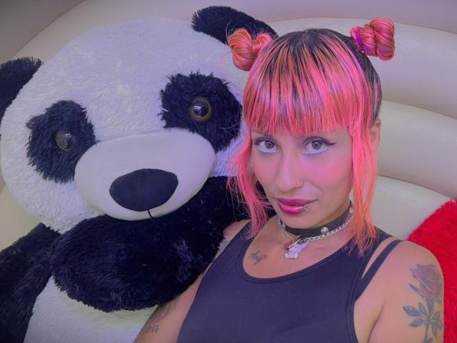 MilaRouge with pink and orange hair in double buns, black tank top, choker, tattoos, and pink lipstick, next to a giant black and white stuffed panda.