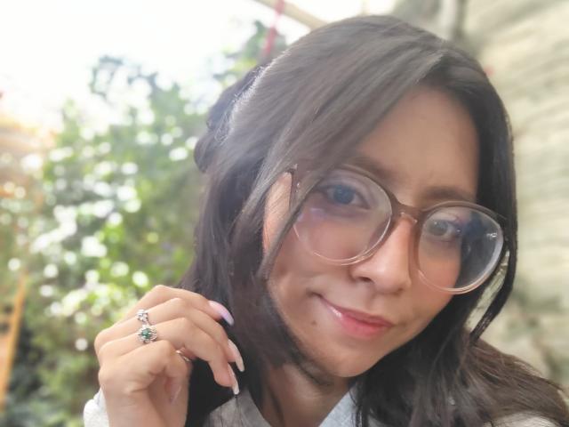 VioletXBloom with round glasses, black hair, light skin, and a subtle smile, showing a ring with a green stone on her finger, slightly blurry background.
