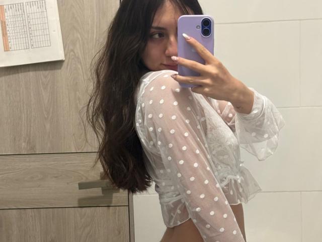 Luluberrie with long brown hair, wearing a sheer white polka dot top, showing her tits in a mirror selfie, holding a purple phone, in a bathroom.