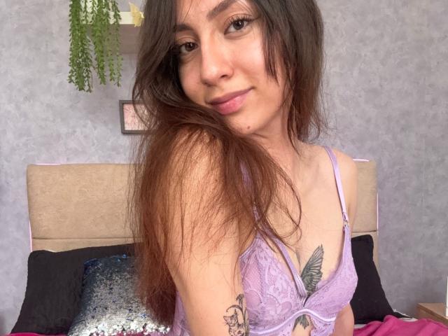 Luluberrie with long brown hair, purple lace bra, small tits, tattoo on right chest, subtle smile, brown eyes, and light skin, sitting on bed with black and sequined pillows.