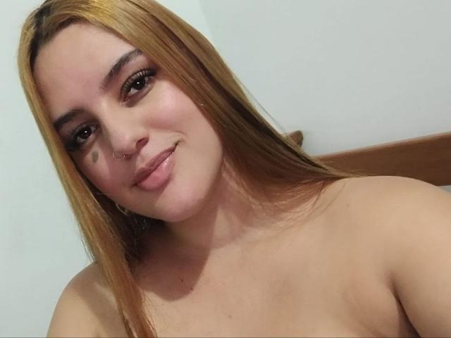 Hailey Tims, com cabelo castanho claro, piercing no nariz e uma pequena tatuagem preta na bochecha, sorrindo com os lábios curvados, sem blusa, mostrando seus ombros e parte superior do peito..