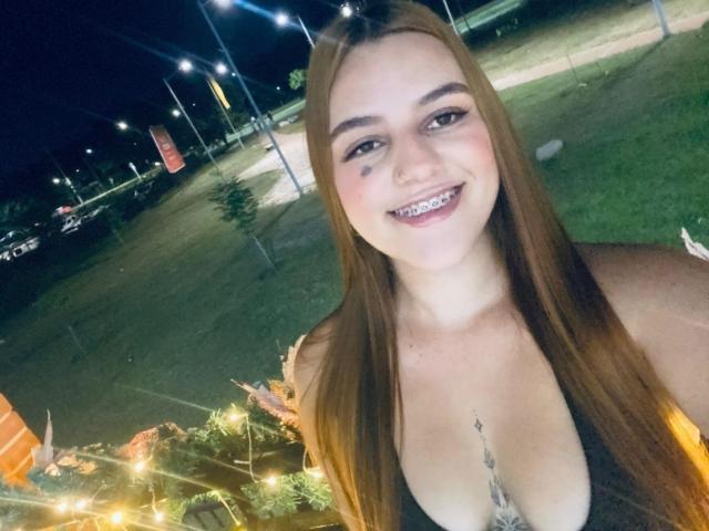 Hailey Tims mit langen, braunen Haaren, lächelnd, mit freiliegenden Zähnen, trägt ein schwarzes Oberteil, das ihre Brüste zeigt, und hat ein kleines Tattoo oberhalb ihrer Brust. Das Foto ist ein Nacht-Selfie..