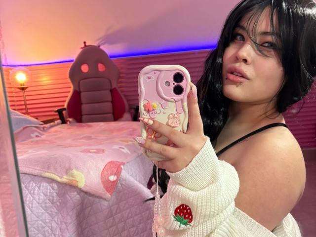 HoneeyBonny with black hair, pink lipstick, and a white sweater with strawberry embroidery, taking a selfie in a pink-themed bedroom, showing her black bra and holding a pink phone.