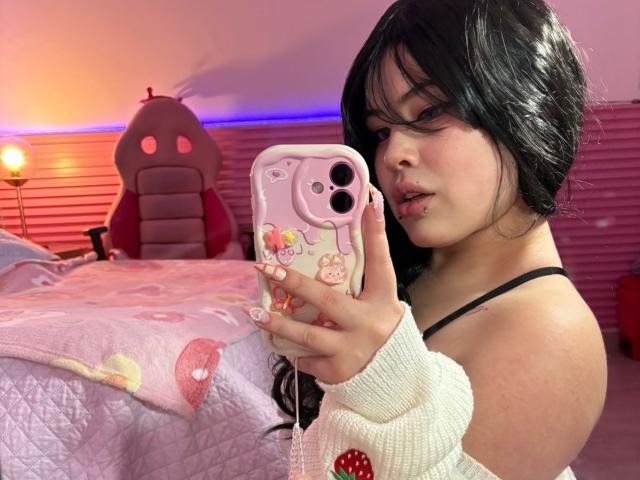HoneeyBonny with black hair, pink phone case, and white sweater, taking a selfie, showing her nose ring and black strap. Her tits are visible, and she's wearing a black strap.