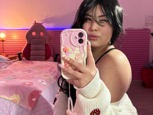 HoneeyBonny with black hair, pouty lips, and a cute pink phone case, taking a selfie in a pink room, wearing a white sweater and black strap. Her tits are slightly visible.