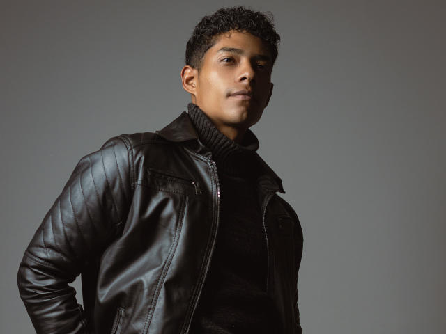 VictoryCassus with short curly black hair, wearing a black quilted leather jacket and black turtleneck, standing against a gray background, looking slightly to the side, with a serious expression.