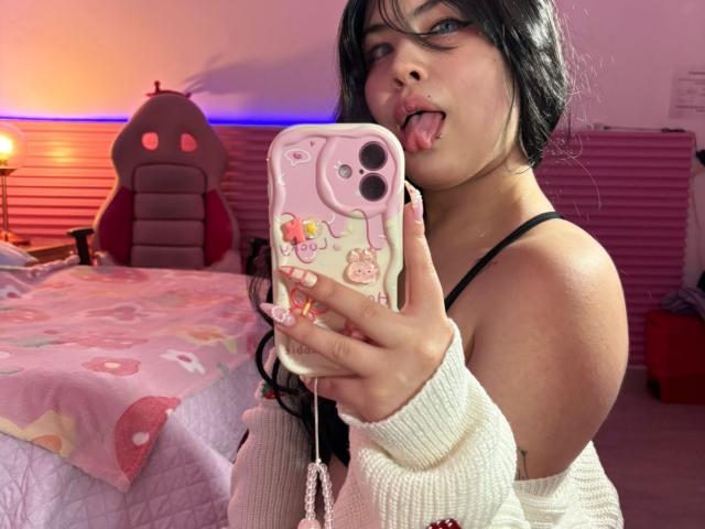 HoneeyBonny with black hair, tongue out, holding pink phone with cute case, wearing white off-shoulder top, exposing black bra strap, and showing pink nails, in a pink bedroom.