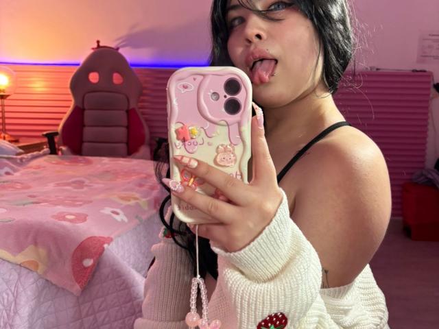 HoneyBonny with black hair, tongue out, pink phone case, white sweater, black strap, holding phone, showing off her tits, pink bed with donut pattern, pink and orange lighting.