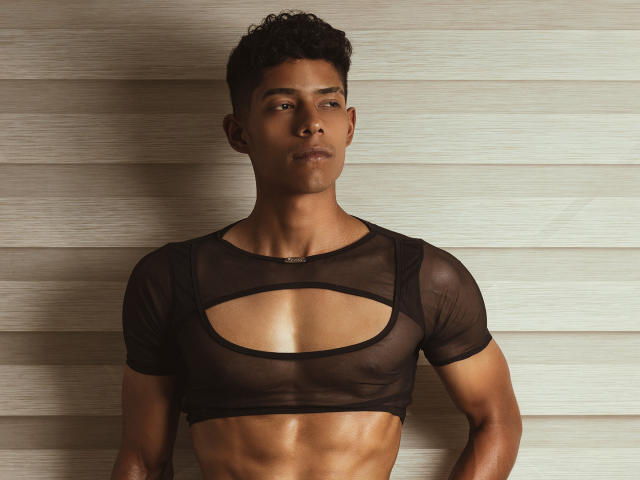 VictoryCassus with curly black hair, brown skin, and a toned, muscular, bare-chested body wearing a sheer black crop top, exposing a cut-out over the chest, looking slightly to the side.