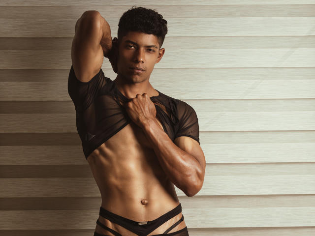 VictoryCassus, muscular and tan, wearing a sheer black top and strappy black underwear, reveals his defined abs and small, hard penis, with one arm raised and a confident look.