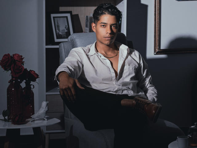 VictoryCassus sits in a chair, wearing a white button-up shirt, black pants, and brown boots, with dark hair, a necklace, and a serious expression, holding his black sock.