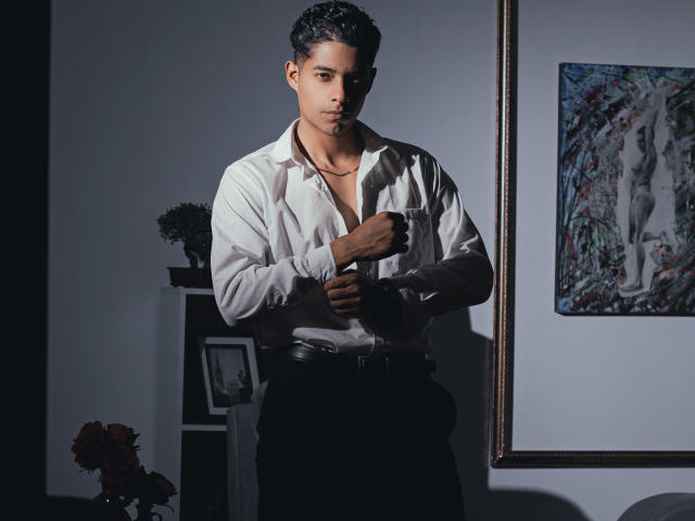 VictoryCassus stands in dim light, wearing a white shirt with chest visible, black pants, and a necklace, hands near shirt, looking serious, dark curly hair, medium skin tone.