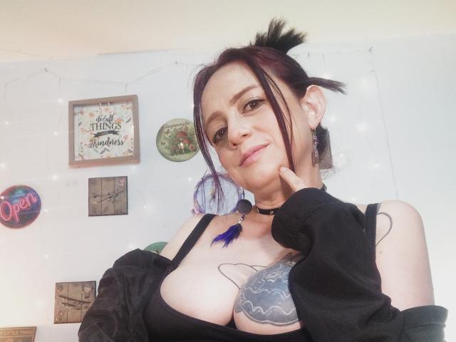 CatalinaMonyque with dark hair in a ponytail, black choker, black bra showing large tits, black jacket, tattoo on right chest, purple hair tie, hand on cheek, smiling.