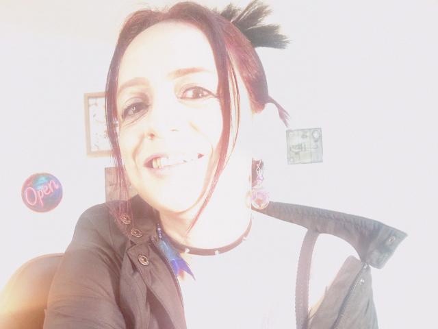 CatalinaMonyque with pale skin, dark hair in pigtails, black choker, smiling with fangs, wearing black shirt, light coming from front, soft glow, slightly overexposed, upper body shot.