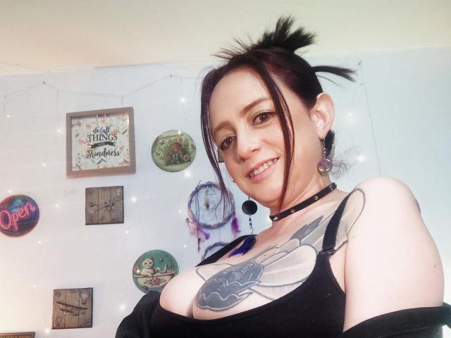 CatalinaMonyque with black hair in a ponytail, wearing a black choker and tank top, showing large tattoos on her chest, smiling at the camera, revealing her tits.