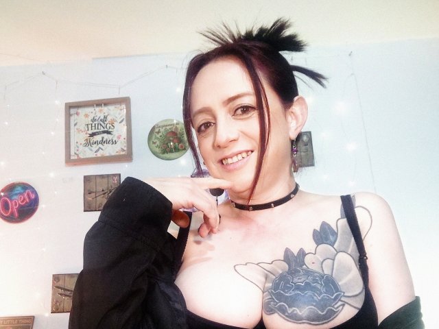 CatalinaMonyque with black hair in a messy bun, smiling, wearing a black choker, showing large tits with a blue flower tattoo, black off-shoulder top, in a bright room.