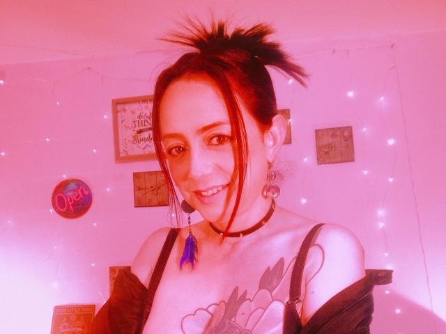 CatalinaMonyque with red-tinted hair in a messy topknot, black choker, and tattoos, smiling, showing her tits in a black strap top, with blue tassel earrings.