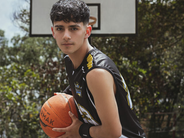 AxellDraven with curly black hair, holding an orange Spalding basketball, wearing a sleeveless black and yellow basketball jersey, standing outdoors, looking slightly to the left, smiling subtly.