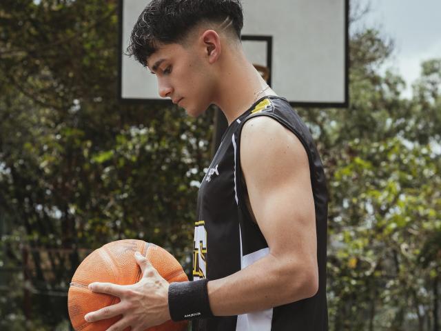 AxellDraven with curly black hair, holding an orange basketball, wearing a black sleeveless sports shirt, white and yellow accents, black wristband, looking down, muscular arms, outdoors.