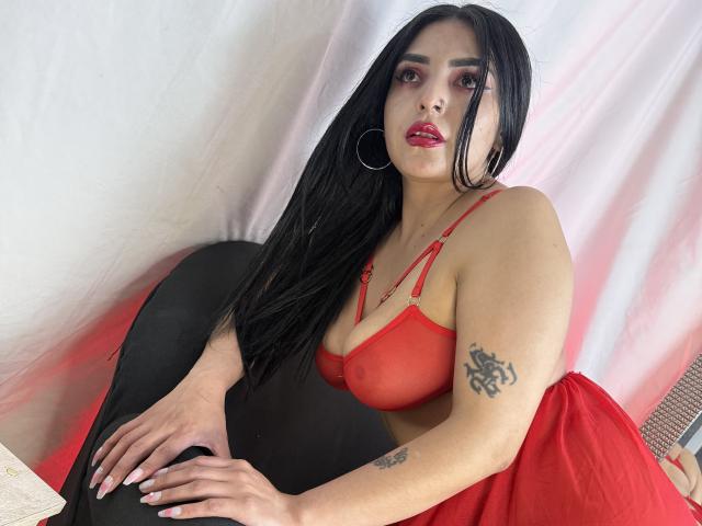CamillaNoir with long black hair, red bra, red lipstick, hoop earrings, tattoo on right arm, red skirt, sitting on black chair, hands on lap, white and pink backdrop.