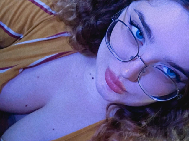 TeasingXDora with curly brown hair, blue eyes, glasses, and a yellow shirt, showing her cleavage and small moles on her chest, looking at the camera with pouty lips.