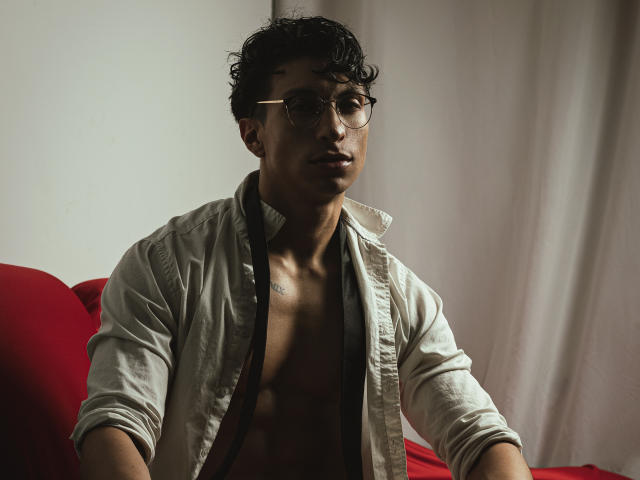 ApoloCrus with curly black hair, glasses, and a partially open gray shirt, revealing a toned chest and dark skin, sitting on a red chair, looking serious.